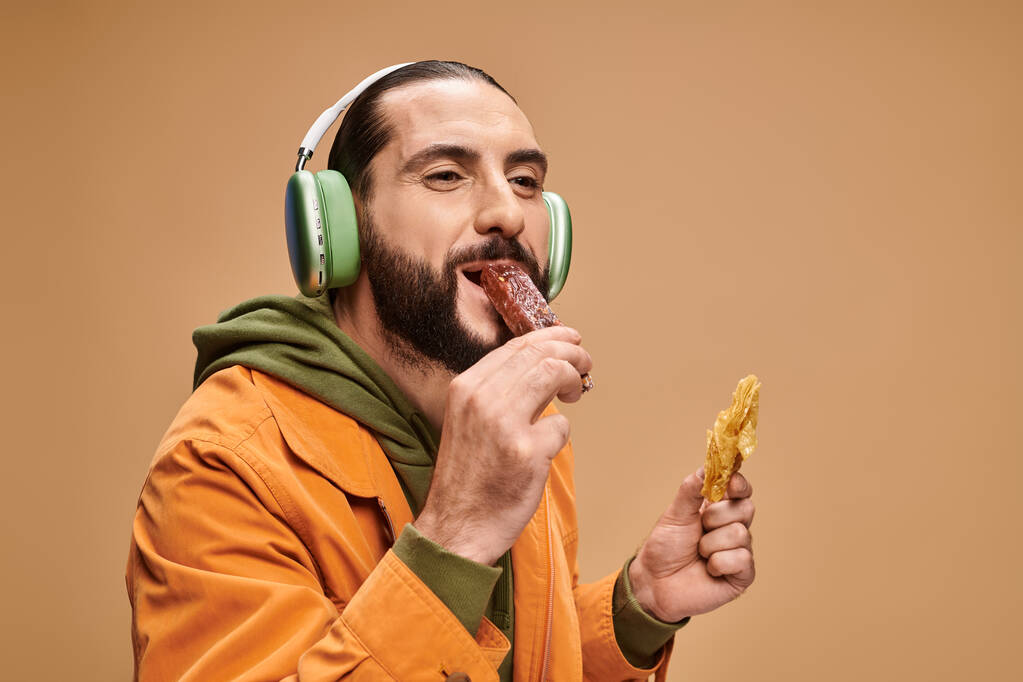 szczęśliwy arabski człowiek w słuchawkach trzyma miód baklava i gryzie churchkhela na beżowym tle - Zdjęcie, obraz