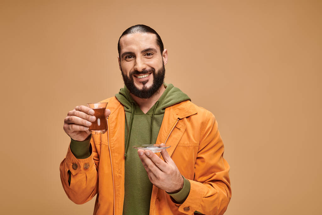 šťastný vousatý muž v ležérní oblečení drží turecký čaj v tradičním skleněném poháru na béžové pozadí - Fotografie, Obrázek