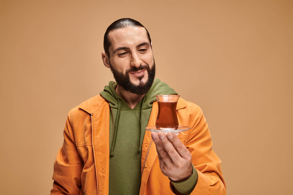 щасливий бородатий чоловік у повсякденному вбранні, дивлячись на турецький чай у скляній чашці на бежевому фоні - Фото, зображення
