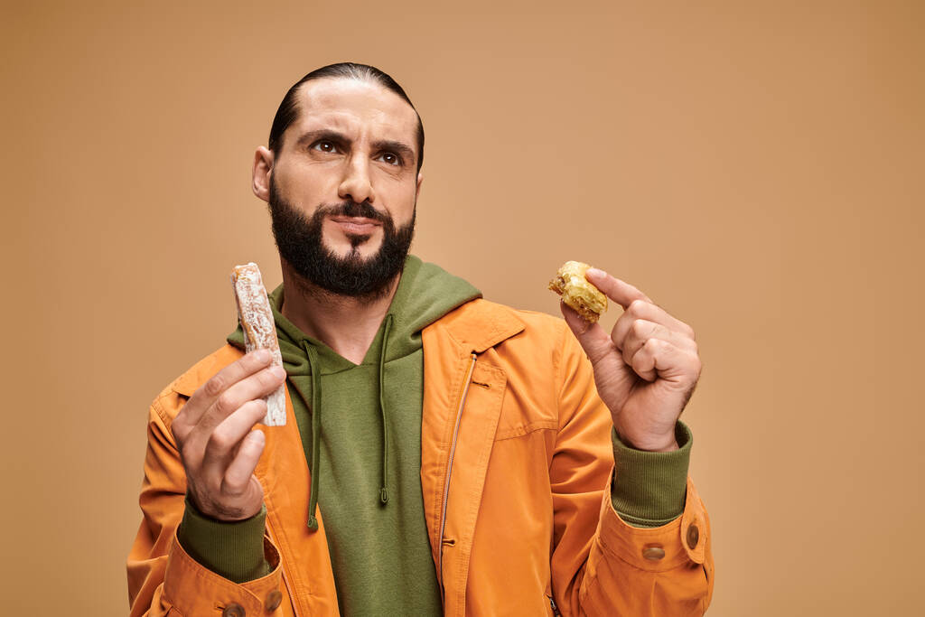 hombre barbudo confundido sosteniendo baklava y cevizli sucuk sobre fondo beige, delicias turcas - Foto, Imagen