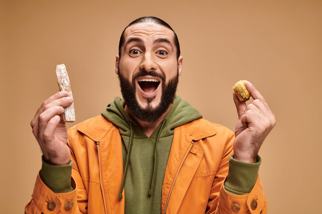 hombre barbudo árabe excitado sosteniendo baklava y cevizli sucuk sobre fondo beige, delicias turcas - Foto, Imagen