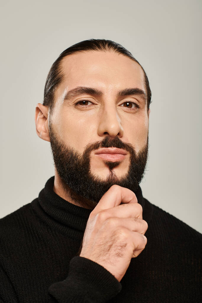 handsome middle eastern man in black turtleneck touching beard on grey background, confidence - Photo, Image