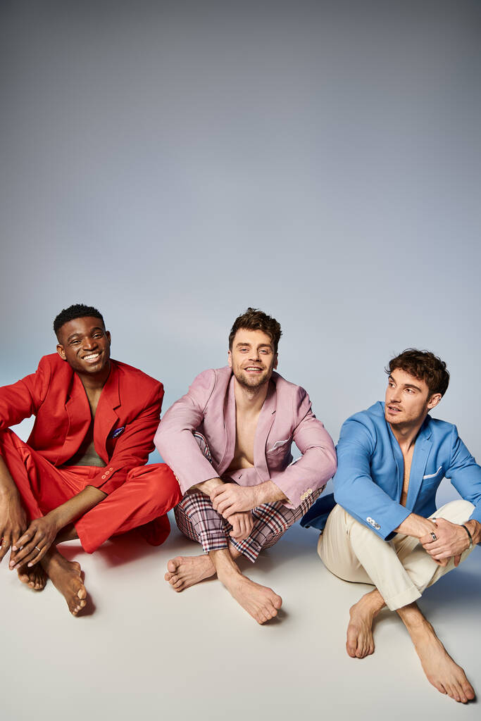 счастливые разнообразные мужчины в ярких костюмах сидят на полу со скрещенными ногами и радостно улыбаются, мода - Фото, изображение