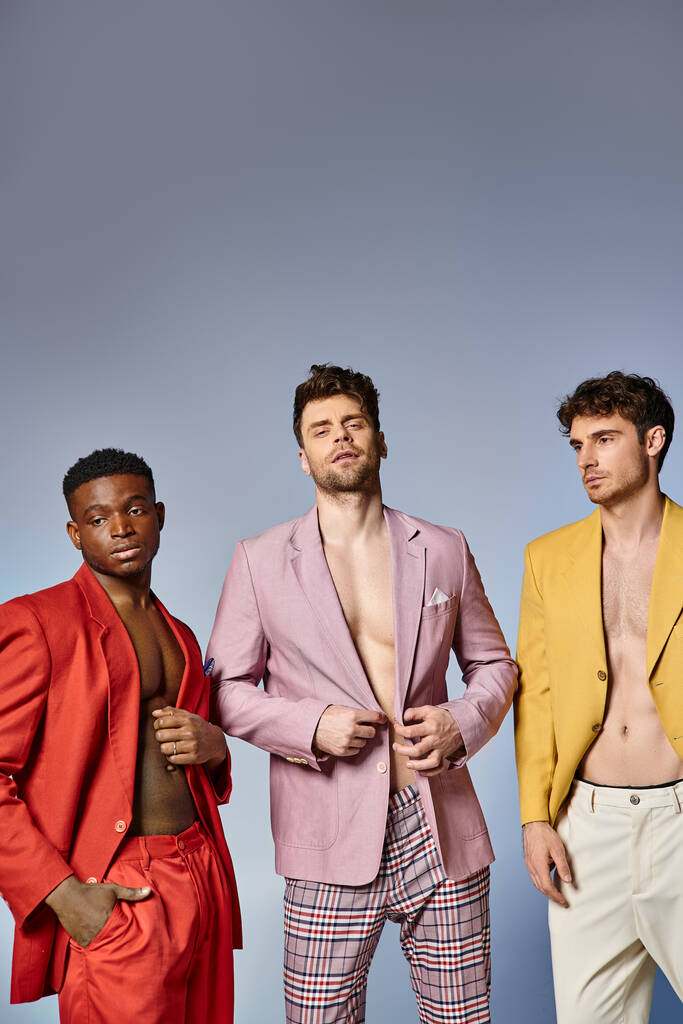 drie verschillende mannen in levendige unbuttoned pakken samen poseren op grijze achtergrond, mode concept - Foto, afbeelding