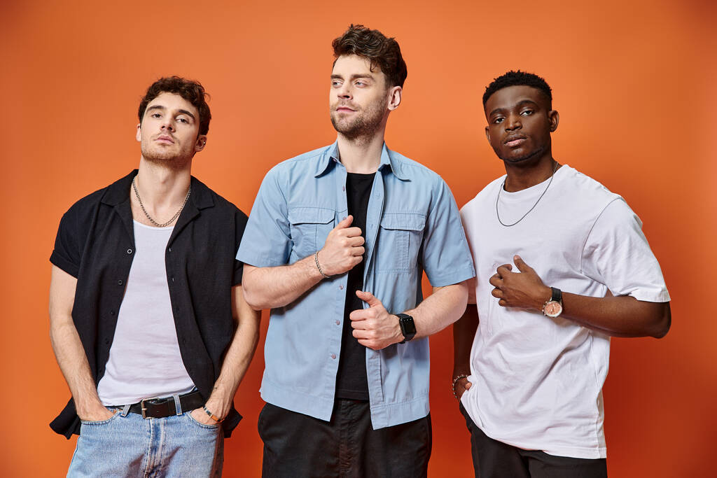 three handsome multicultural friends in casual urban outfits posing together on orange backdrop - Photo, Image