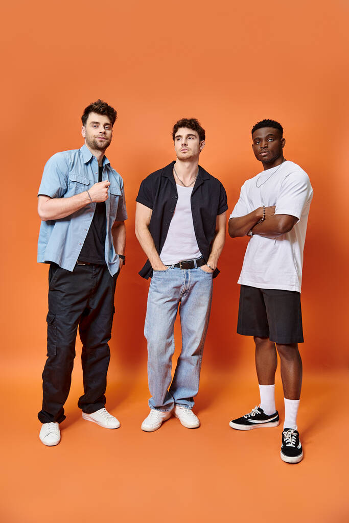 drei attraktive multikulturelle Freunde in lässigen urbanen Outfits posieren gemeinsam vor orangefarbener Kulisse - Foto, Bild