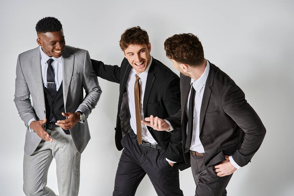 jóvenes felices amigos multiculturales en los negocios elegantes trajes sonriendo alegremente sobre fondo gris - Foto, Imagen