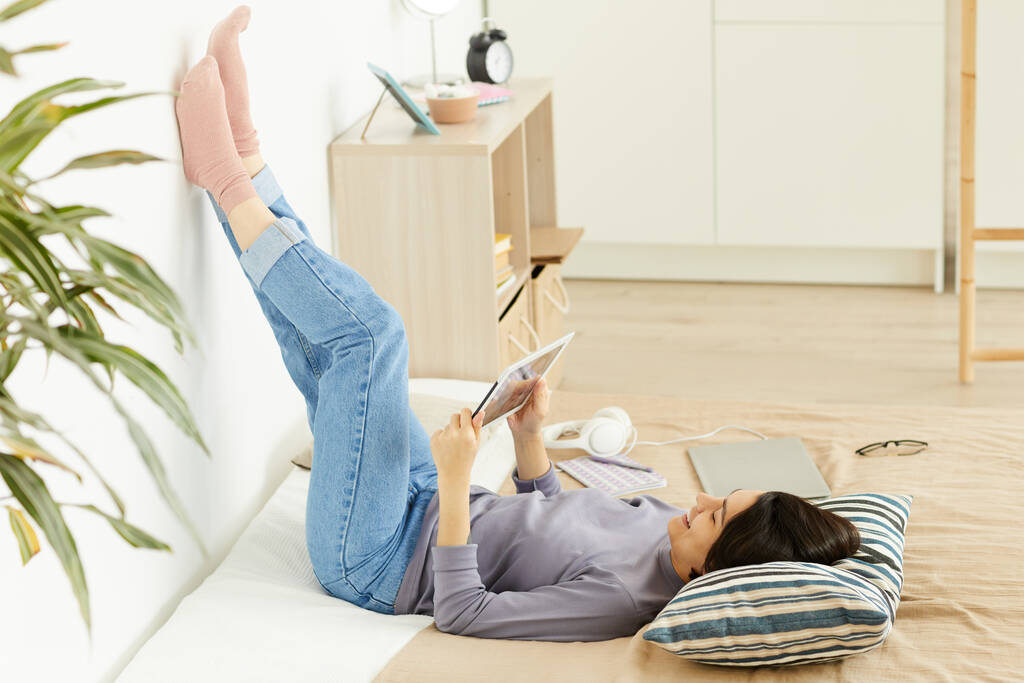 Веселая привлекательная молодая женщина в джинсах, лежащая на кровати и опирающаяся на стену, используя планшет и проводя время в социальных сетях - Фото, изображение