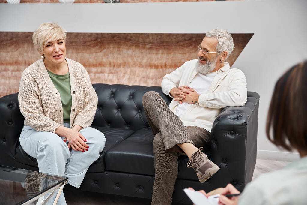 happy middle aged couple sitting on leather and smiling with psychologist during consultation - Photo, Image