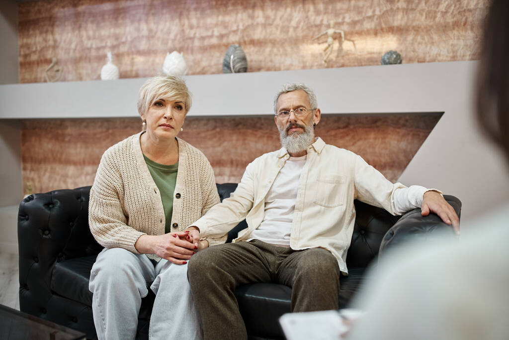 εξυπηρετικό μεσήλικο ζευγάρι κάθεται σε δερμάτινο καναπέ και ψάχνει σε ψυχολόγο κατά τη διάρκεια της διαβούλευσης - Φωτογραφία, εικόνα