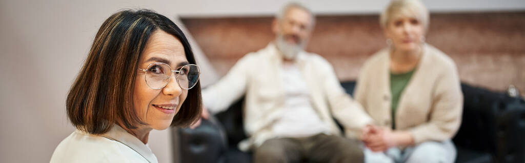 баннер, психолог средних лет в очках, смотрящий на камеру рядом с супружеской парой на заднем плане - Фото, изображение