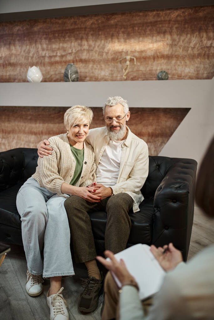 щаслива подружня пара середнього віку обіймає і сидить на шкіряному дивані під час сеансу сімейної терапії - Фото, зображення