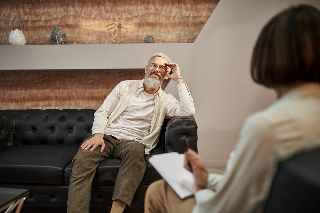 веселий бородатий чоловік середнього віку з татуюванням сидить на шкіряному дивані і дивиться на психолога - Фото, зображення