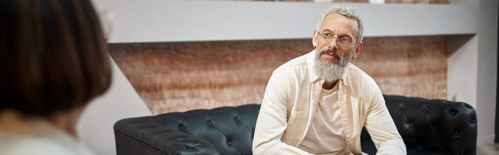бородатый мужчина среднего возраста с татуировкой, сидящий на кожаном диване и слушающий психолога, баннер - Фото, изображение
