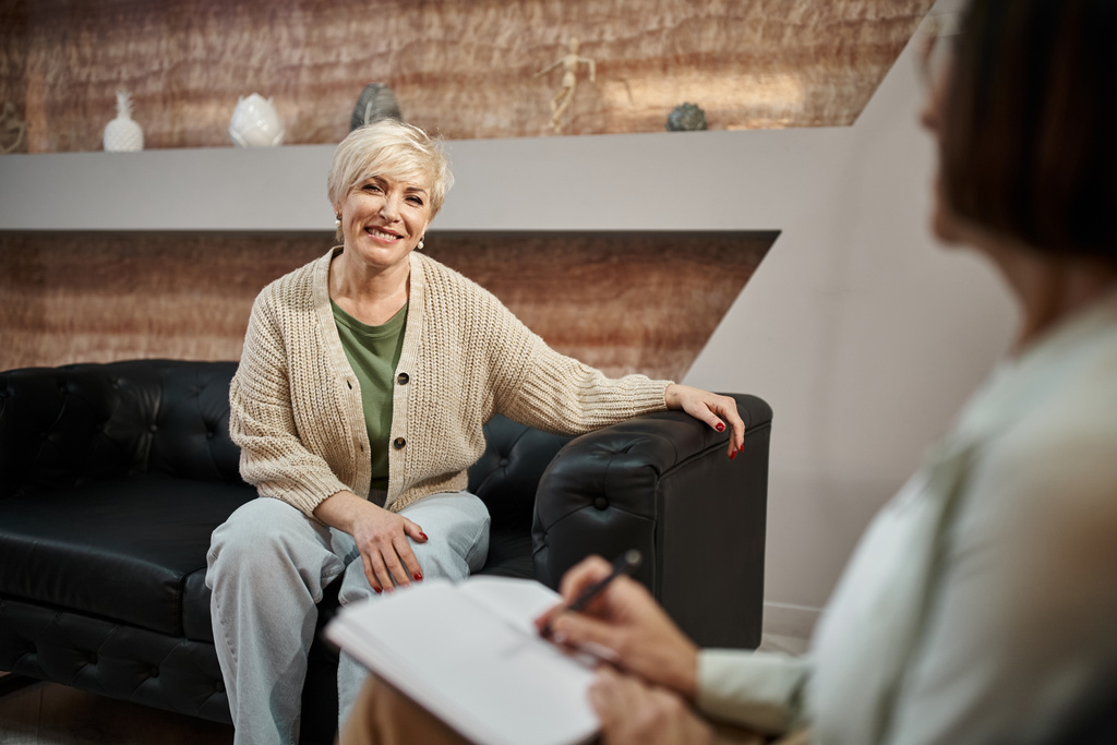 χαρούμενη ξανθιά μεσήλικη γυναίκα κάθεται σε δερμάτινο καναπέ και ψάχνει σε ψυχολόγο σε συνεδρία - Φωτογραφία, εικόνα