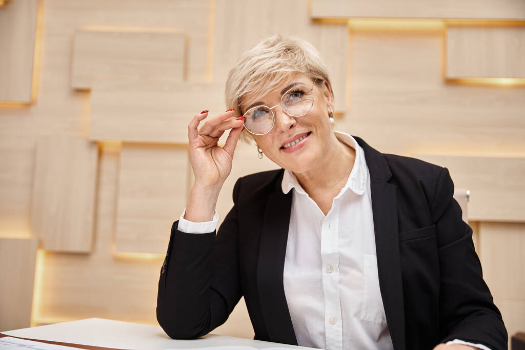 szczęśliwy blondynka w średnim wieku pośrednik w okularach pracy w biurze nieruchomości, patrząc na kamery - Zdjęcie, obraz
