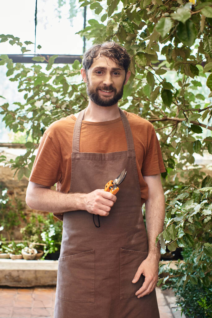 gelukkig tuinman in linnen schort houden tuinieren schaar in kas, poseren met de hand in zak - Foto, afbeelding