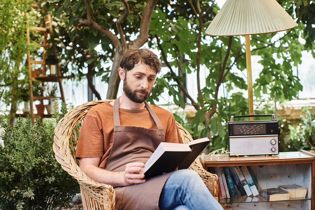 hermoso jardinero barbudo en delantal de lino lectura libro y sentado alrededor de las plantas en invernadero - Foto, imagen
