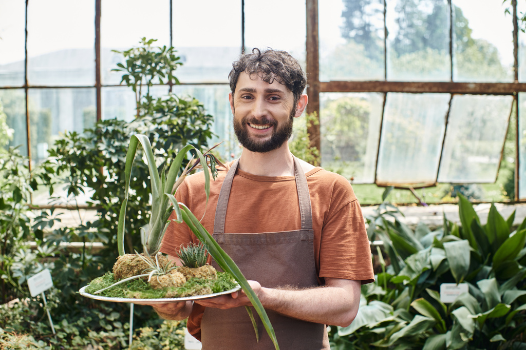 веселий і бородатий садівник в фартусі дивиться на камеру і тримає зелену рослину в теплиці - Фото, зображення