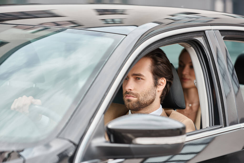 αυτοπεποίθηση γενειοφόρος άνδρας οδήγηση πολυτελές αυτοκίνητο με businesswoman στο πίσω κάθισμα σε θολή φόντο - Φωτογραφία, εικόνα