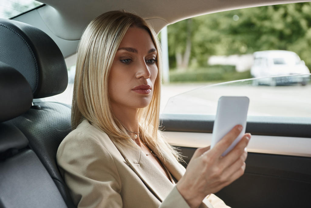 ξανθιά επιχειρηματίας σε κομψό επίσημη φθορά περιήγηση στο διαδίκτυο στο κινητό τηλέφωνο, ενώ κάθεται στο αυτοκίνητο - Φωτογραφία, εικόνα