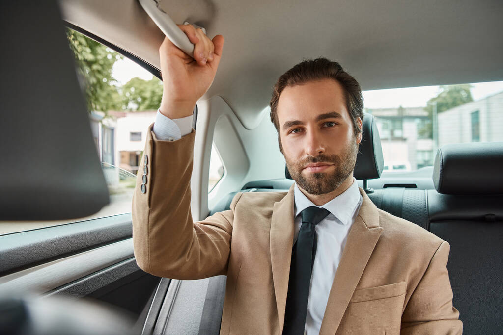 уверенный и красивый бизнесмен в бежевом элегантном костюме смотрит на камеру в роскошном автомобиле на улице - Фото, изображение