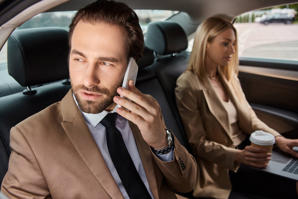 businessman in formal wear talking on smartphone near woman with coffee to go and laptop in car - Photo, Image