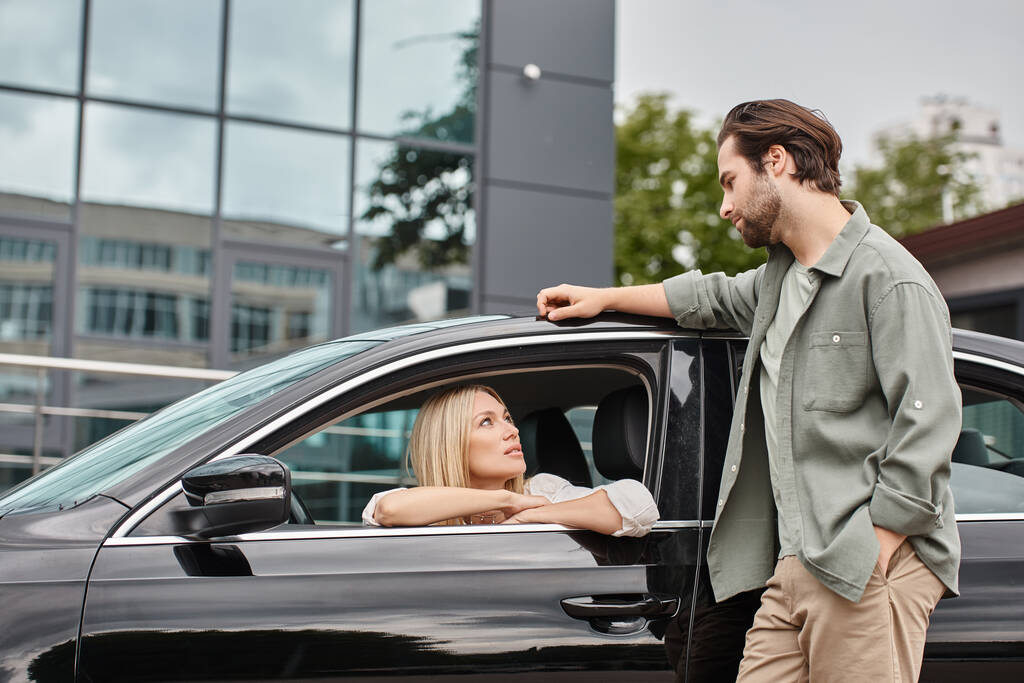 przystojny i stylowy mężczyzna stoi w pobliżu atrakcyjne blondynka siedzi w nowoczesnym samochodzie, uczucie - Zdjęcie, obraz