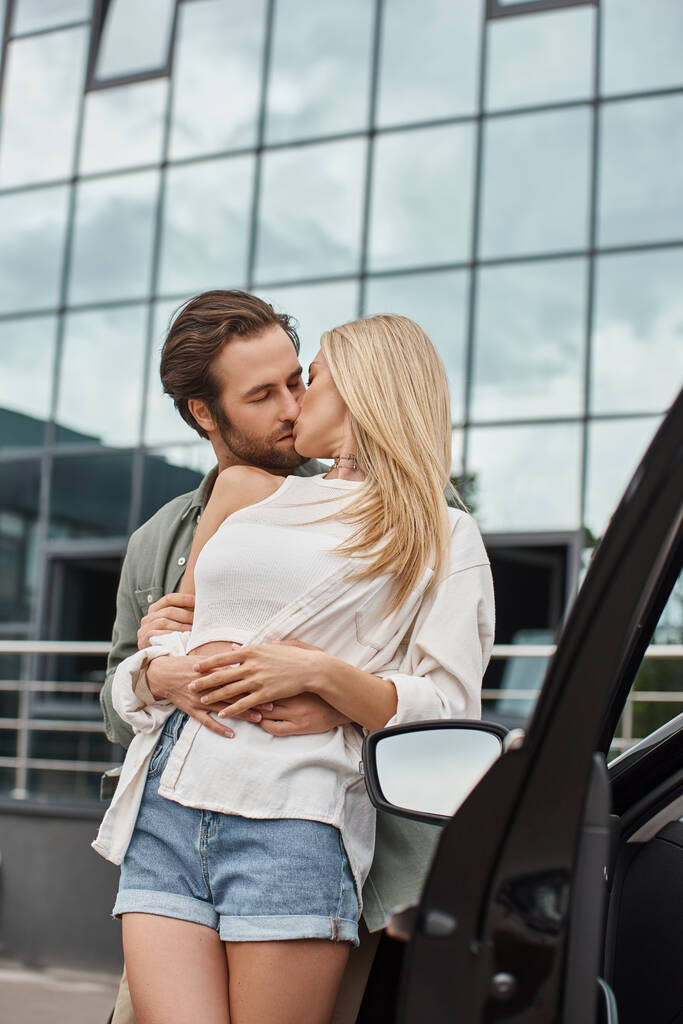 elegante hombre guapo abrazando y besando novia rubia cerca de coche moderno en la calle urbana - Foto, imagen