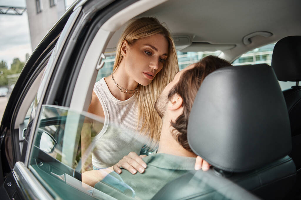 привлекательная блондинка соблазняет мужчину, сидящего в современном автомобиле на городской улице, романтические любовные свидания - Фото, изображение