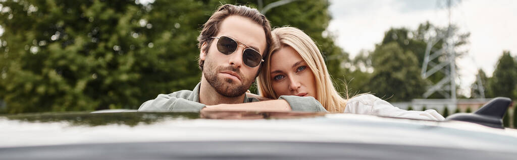 όμορφος άνδρας με γυαλιά ηλίου και αισθησιακή ξανθιά γυναίκα κοιτάζοντας κάμερα κοντά στο αυτοκίνητο στο δρόμο, πανό - Φωτογραφία, εικόνα