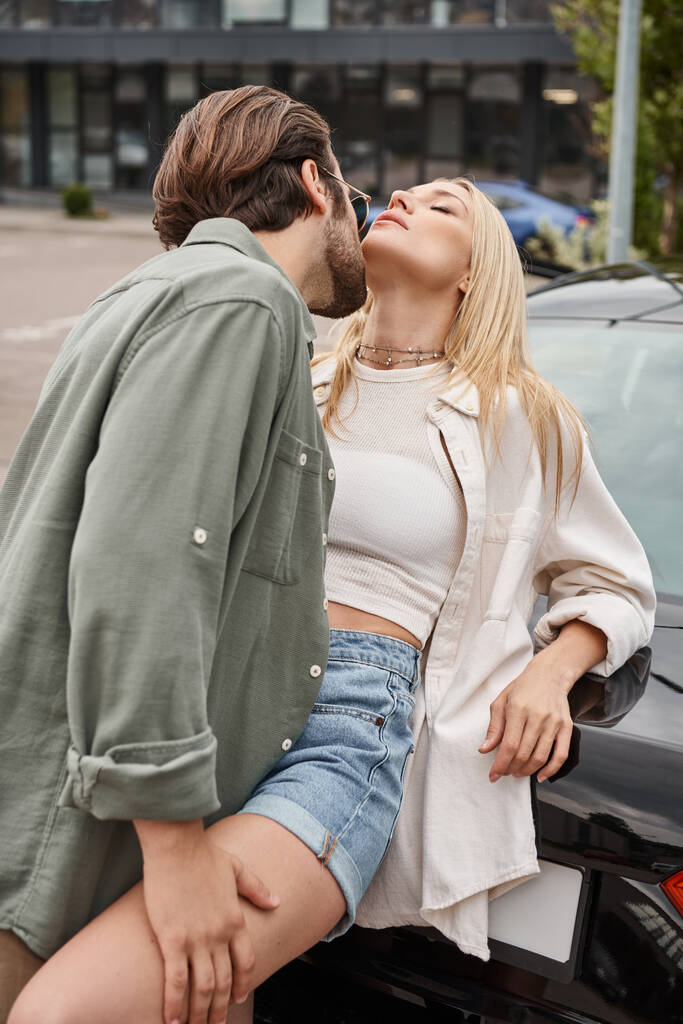 mężczyzna w stylowe casual ubrania i okulary uwodzenie zmysłowy blondynka kobieta w pobliżu samochodu na ulicy - Zdjęcie, obraz
