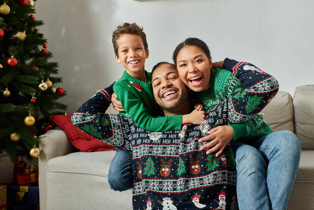 familia afroamericana feliz en suéteres acogedores abrazándose calurosamente y sonriendo a la cámara, Navidad - Foto, imagen