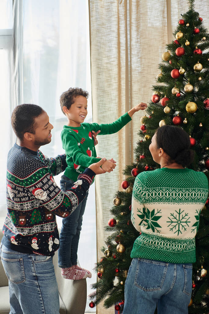 verticaal schot van Afrikaanse Amerikaanse familie glimlachen vrolijk naar elkaar versieren kerstboom - Foto, afbeelding