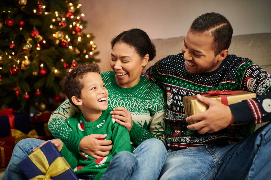 счастливая африканская американская семья сидит рядом с елкой с гирляндами и улыбается друг другу - Фото, изображение