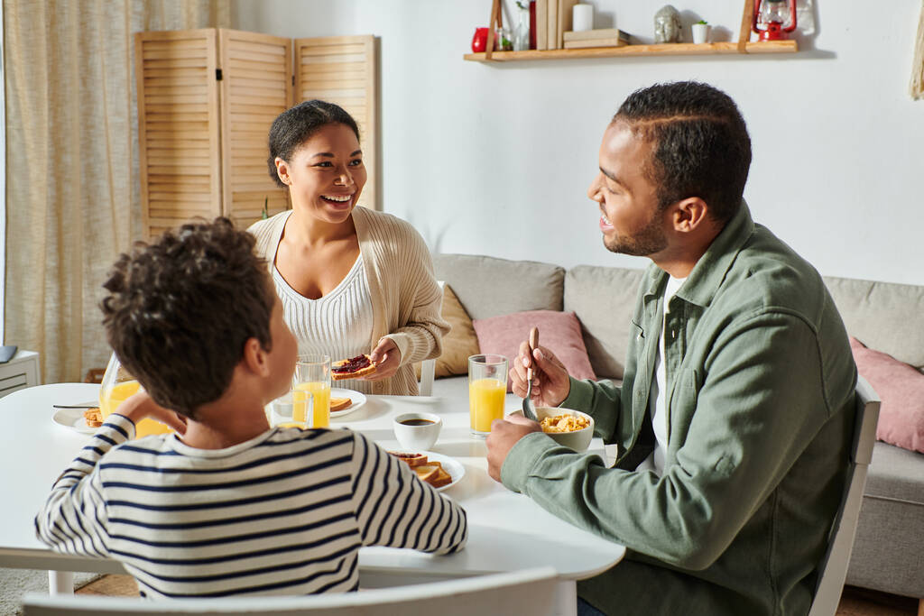 familia afroamericana feliz en trajes casuales sonriendo amorosamente el uno al otro durante el desayuno - Foto, Imagen