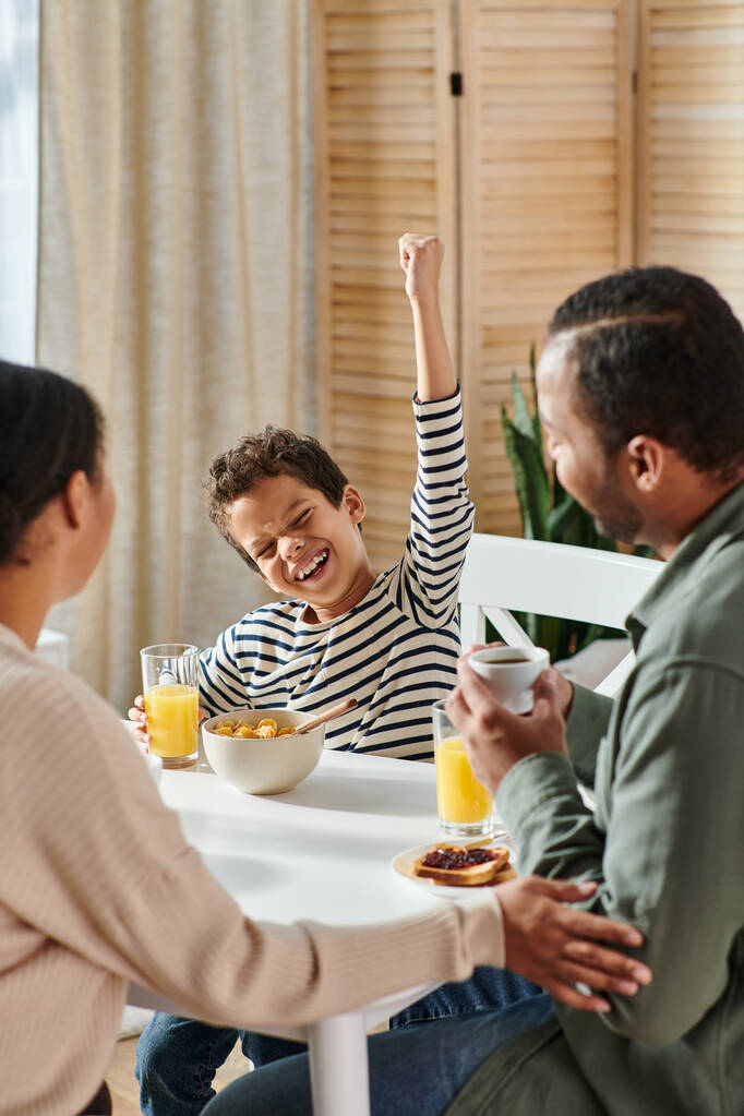 vertikale Aufnahme eines kleinen afrikanisch-amerikanischen Jungen, der beim Frühstück jubelt, während seine Eltern ihn anschauen - Foto, Bild