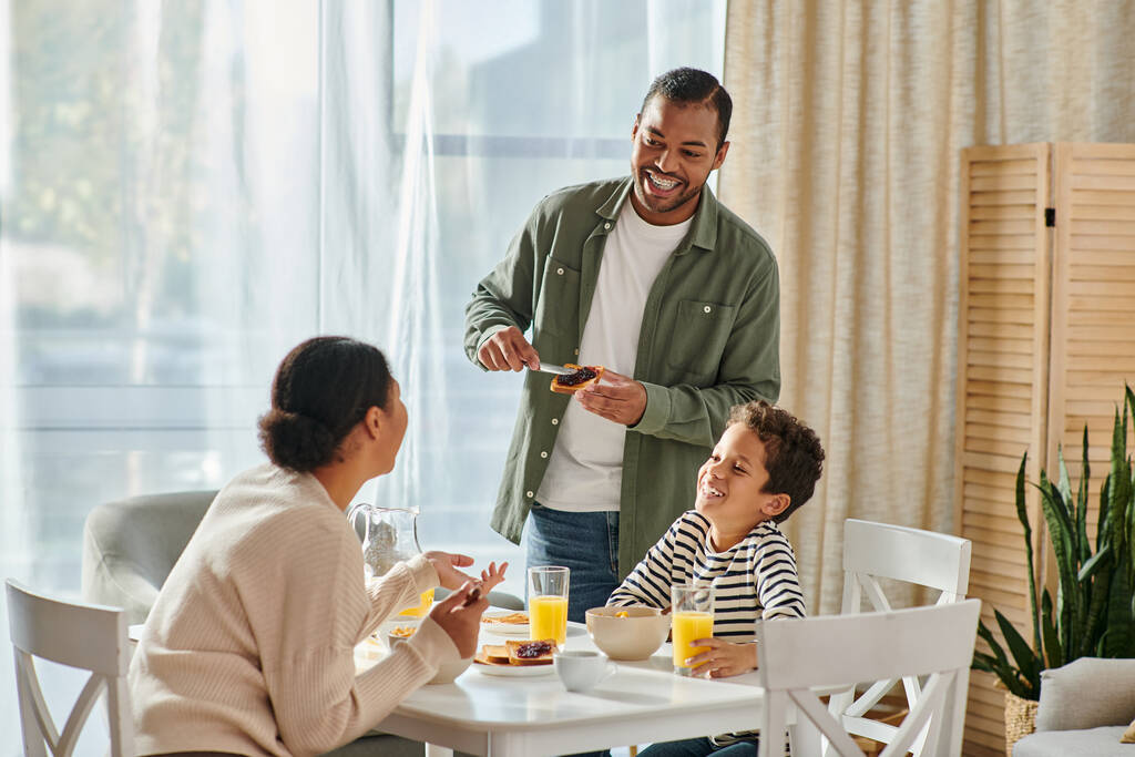 veselý africký Američan roztahuje marmeládu na toast a dívá se na svou ženu a syna u snídaně - Fotografie, Obrázek