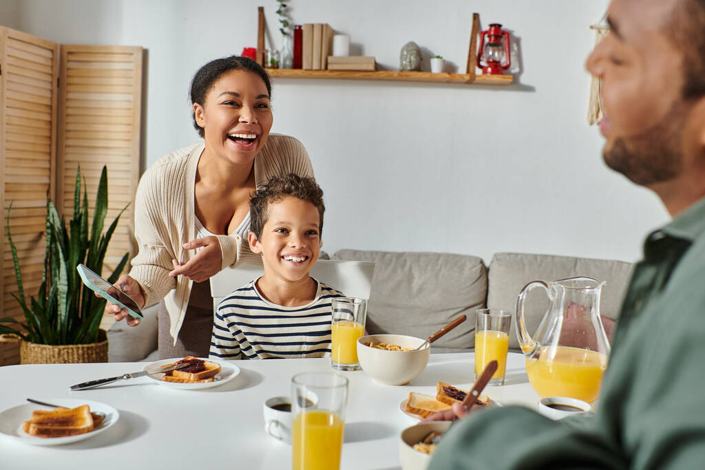 focus on cheerful african american mother and son in homewear smiling at blurred father at breakfast - Photo, Image