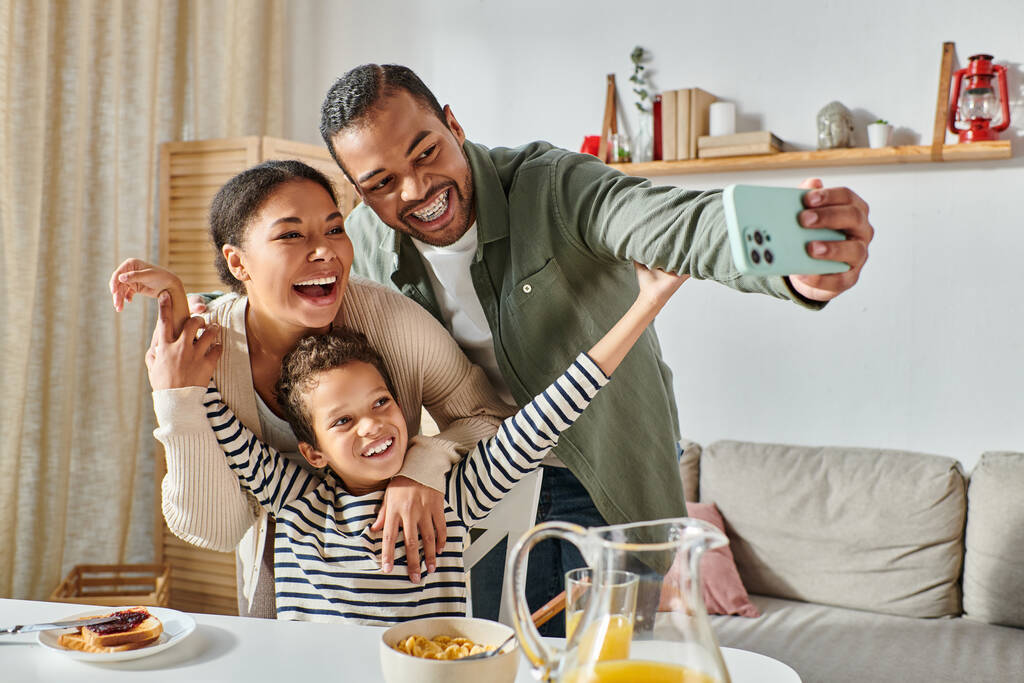 nowoczesna, wesoła afrykańska rodzina robi sobie selfie przy stole śniadaniowym i uśmiecha się do kamery - Zdjęcie, obraz