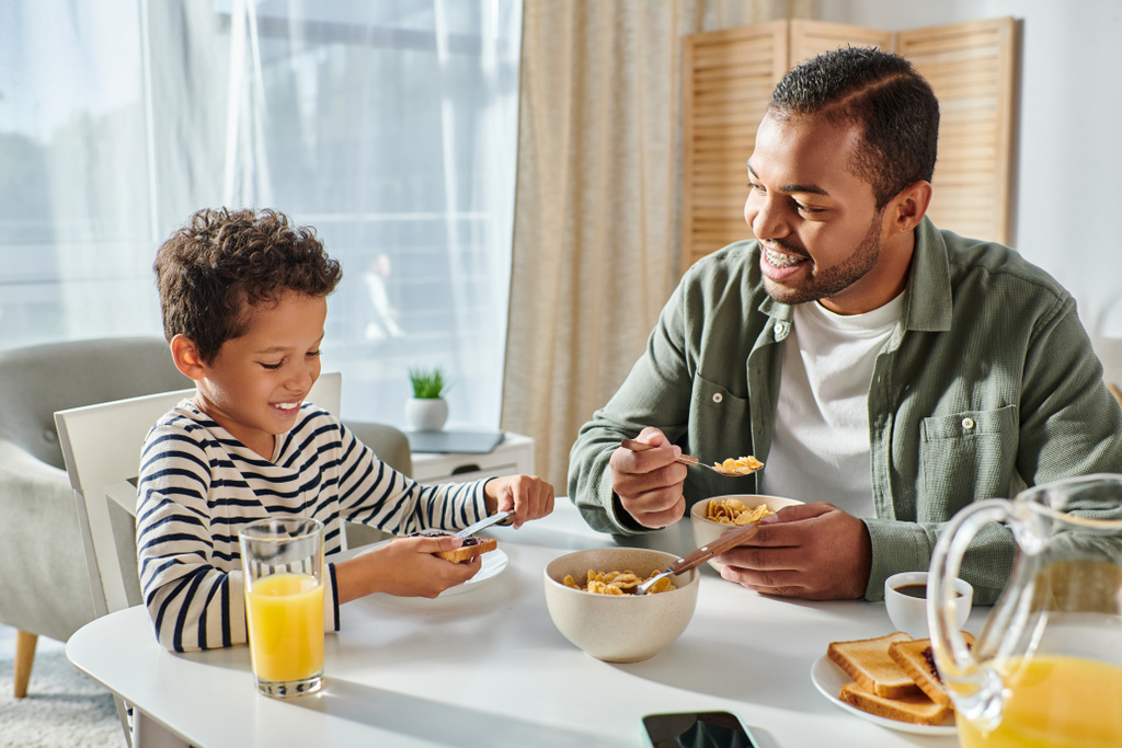 iloinen komea afrikkalainen amerikkalainen isä etsii onnellisesti hänen poikansa syö aamiaista pöydässä - Valokuva, kuva