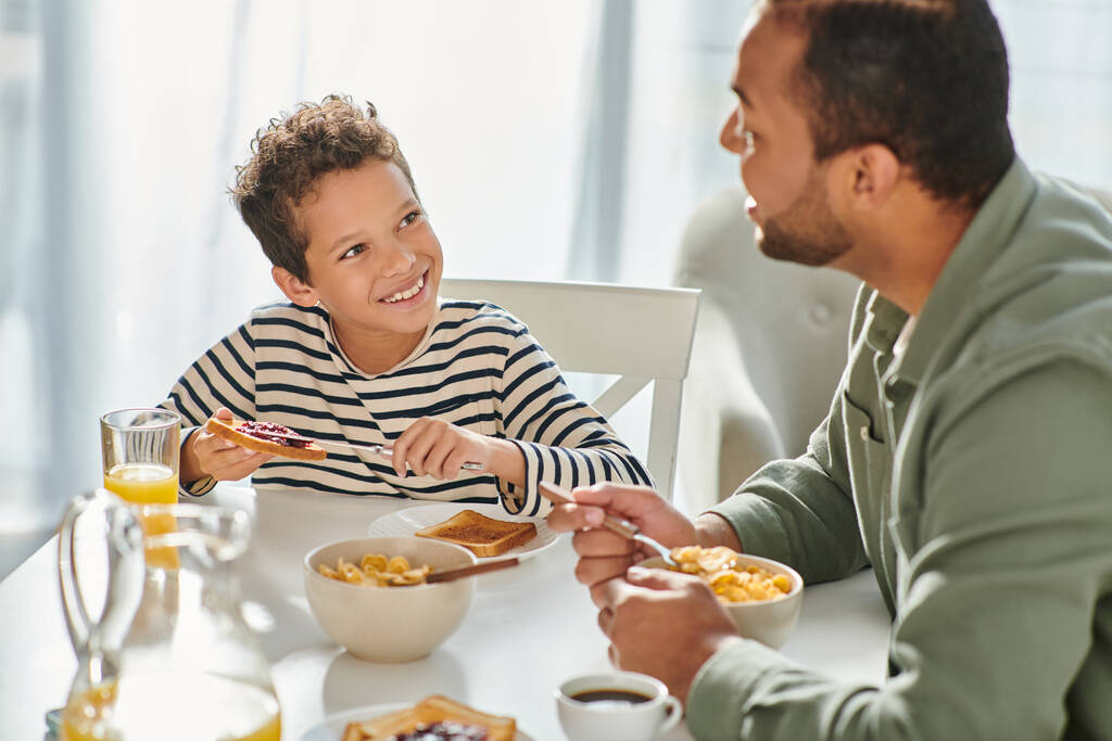 joyeux père afro-américain et fils en vêtements de maison petit déjeuner et souriant à l'autre - Photo, image