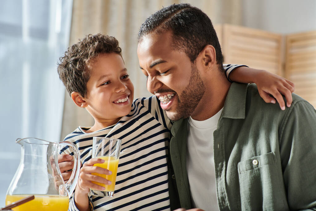 fröhlicher afrikanisch-amerikanischer Junge mit Orangensaft umarmt seinen Vater an der Schulter am Frühstückstisch - Foto, Bild