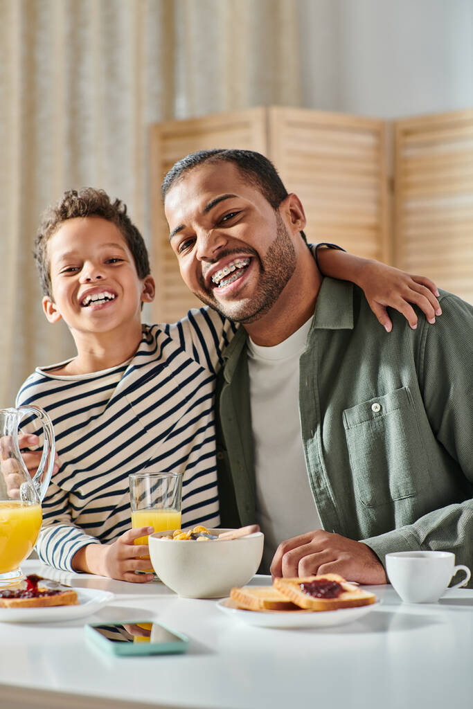 tiro vertical de alegre afro-americano pai e filho abraçando no café da manhã e sorrindo para a câmera - Foto, Imagem