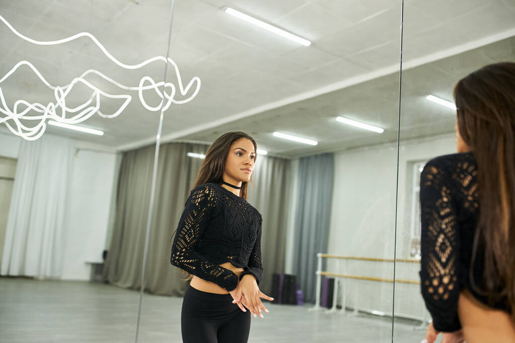 Мистецька афроамериканська танцівниця в чорному одязі дивиться у дзеркало і тренується в танцювальному залі - Фото, зображення