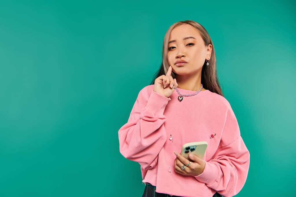 Pembe kazaklı, düşünceli genç Asyalı kadının portresi turkuaz arka planda akıllı telefon kullanıyor. - Fotoğraf, Görsel