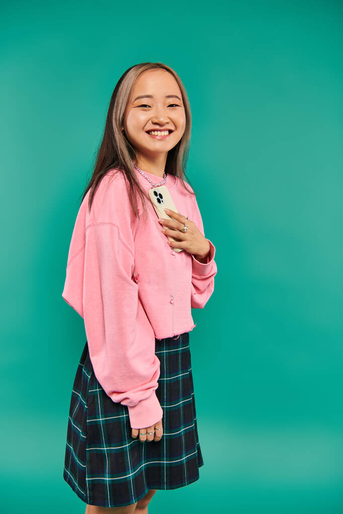 muotokuva positiivinen nuori aasialainen nainen vaaleanpunainen huppari ja ruudullinen hame tilalla älypuhelin sininen - Valokuva, kuva