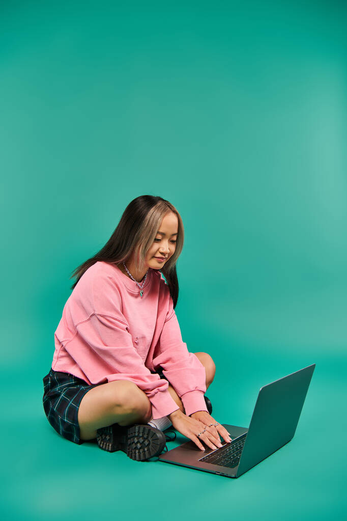 young asian woman in sweatshirt and plaid skirt sitting and using laptop on turquoise backdrop - Photo, Image