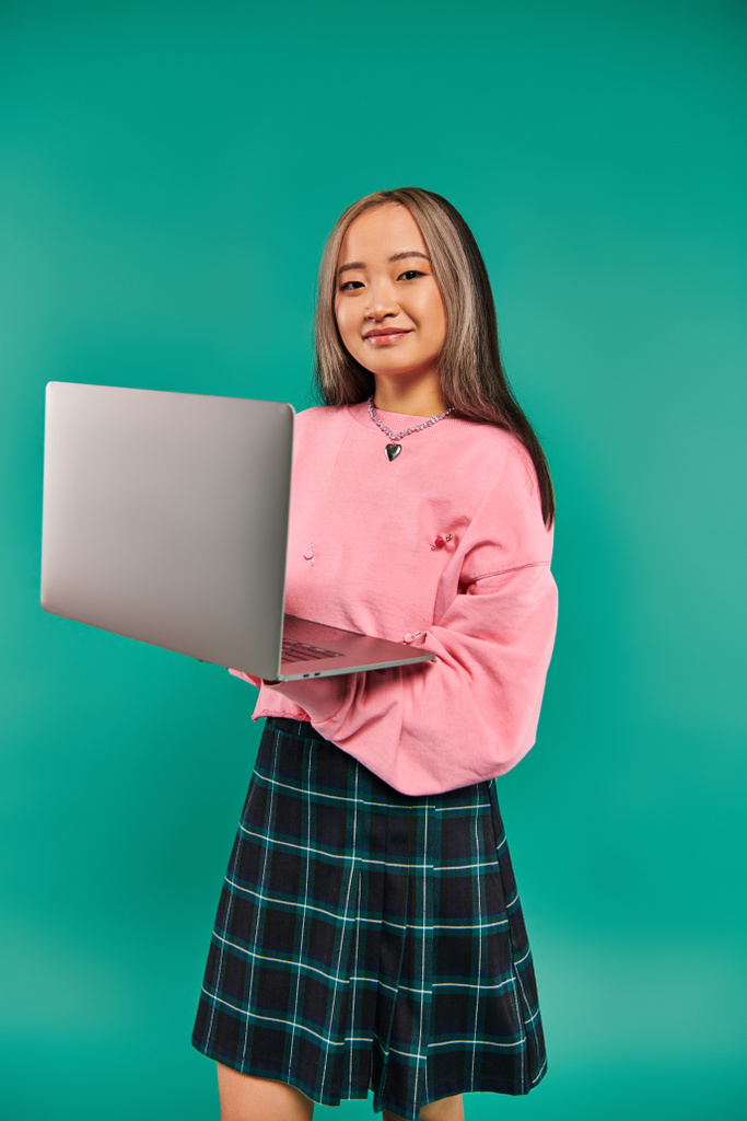 щаслива азіатська дівчина в рожевому светрі, використовуючи ноутбук, стоячи на бірюзовому фоні, мережа - Фото, зображення