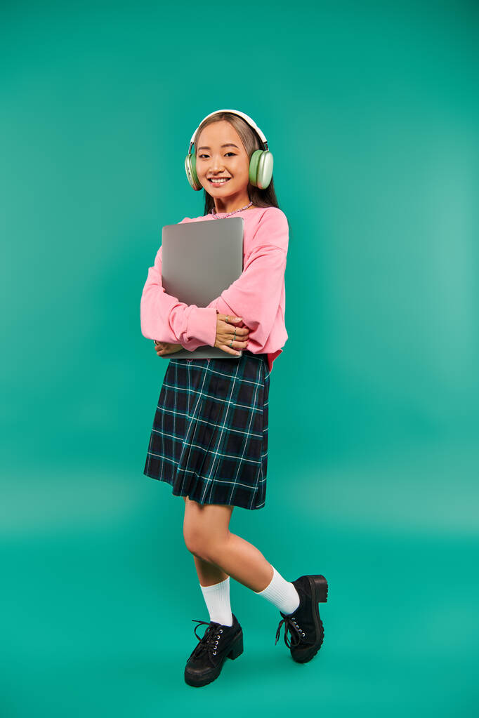 alegre asiático chica en inalámbrico auriculares celebración de portátil y de pie en turquesa telón de fondo - Foto, Imagen